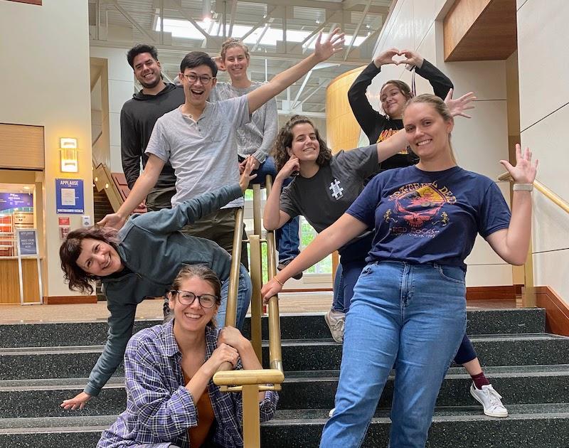 8 people standing on a stairwell holding on to the railing smiling with their arms stretched outward