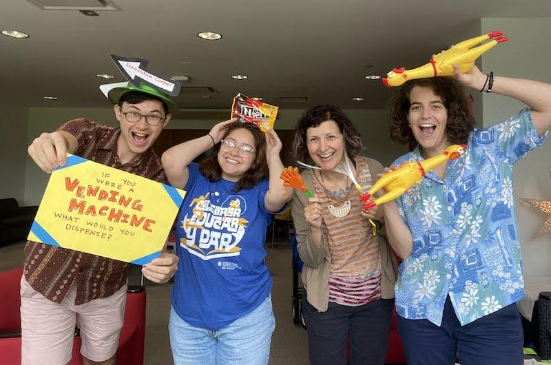 Four people standing in a line smiling holding odd items: a sign that says, "If you were a vending machine, what would you vend?", a bag of M&Ms, a plastic hand clapper, and two rubber chickens