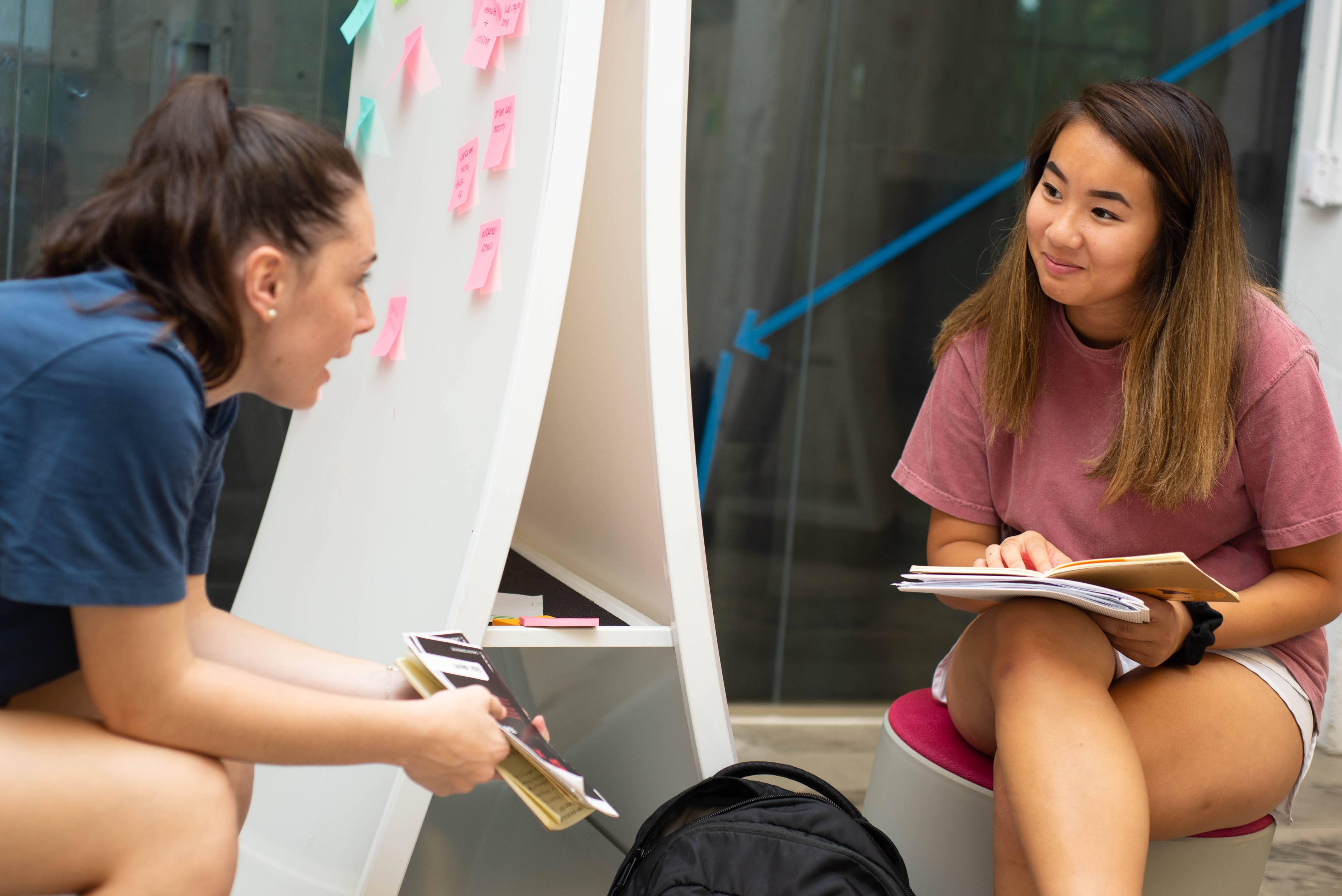 Two students sitting down facing each other. One is talking the other is listening while smiling