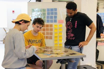 One student standing and two students sitting around a table looking a photos together on the table